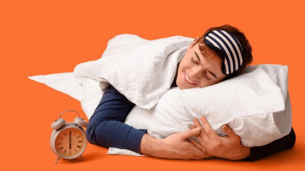 Young man asleep with a striped sleep mask and pillow on an orange background, clock nearby.