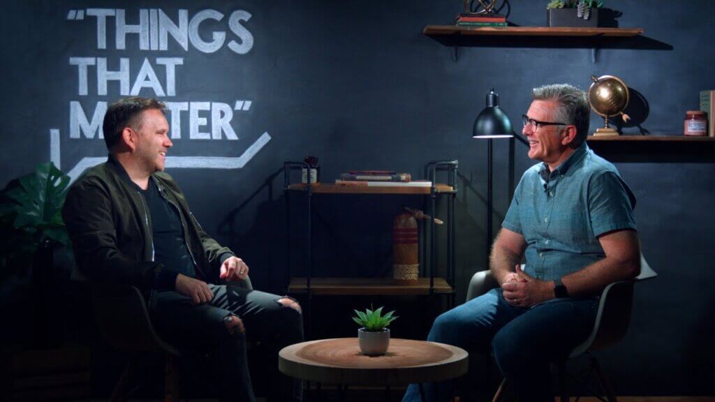 Two men sit across from each other in a studio interview setup, with a round table and a small plant between them, and a wall reading “THINGS THAT MATTER.”