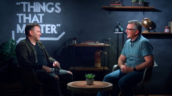 Two men sit across from each other in a studio interview setup, with a round table and a small plant between them, and a wall reading “THINGS THAT MATTER.”