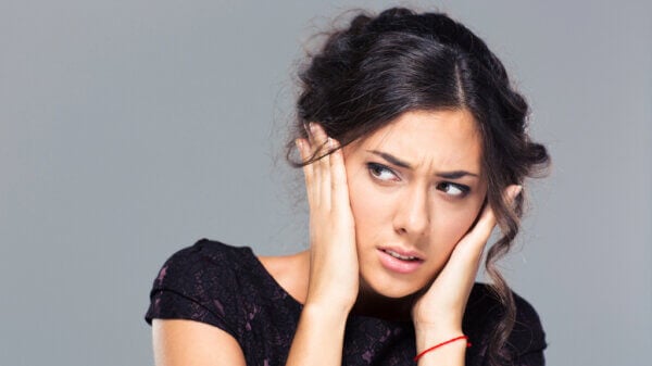 Woman with dark hair holding her ears, showing a worried expression against a gray background.