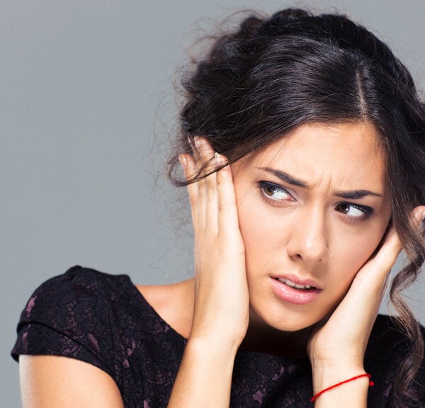 Woman with dark hair holding her ears, showing a worried expression against a gray background.