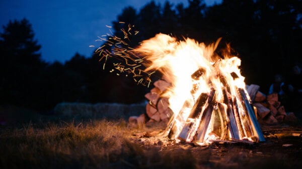 Large bonfire with stacked logs burning at night, sparks flying as flames rise against a dark forest backdrop.