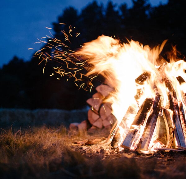 Large bonfire with stacked logs burning at night, sparks flying as flames rise against a dark forest backdrop.